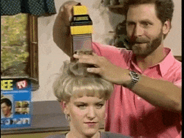 Slow motion infomercial features a man in a pink collared shirt looking mesmerized as he uses a handheld Flowbee hair vacuum to cut and suck the hair of a woman with a sort of bowl cut hairstyle. The woman gives a sharp side eyed expression like she’s checking to see if anyone’s laughing at her. 