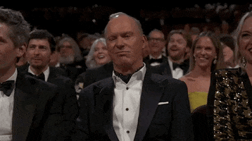 Oscars 2024 GIF. Michael Keaton, seated at the Oscars, dramatically sneers, smirking with a sarcastic chuckle.