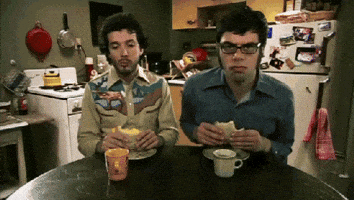 TV gif. Bret McKenzie and Jemaine Clement in Flight of the Conchords  sway rhythmically as they sit at a table eating sandwiches.