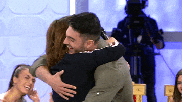 Video gif. Actor Toñi Moreno embraces a woman and picks her up in what appears to be a TV set with other people seated in chairs and watching on with big smiles. 