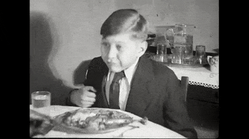 Video gif. Black and white vintage footage of a boy in a suit seated at a table, chewing food happily.