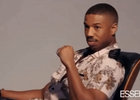 Celebrity gif. Michael B Jordan leans casually in a chair and gradually smiles, then winks.