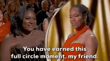 Oscars 2024 gif. Split screen of Regina King adorning a bright red dress onstage and addressing Danielle Brooks proudly by saying, "You have earned this full circle moment, my friend." Brooks wears a sleeveless black dress and smiles at King admiringly before closing her eyes and pumping her fist happily. 