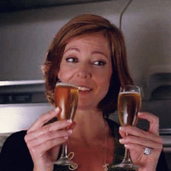 Movie gif. As a flight passenger, Allison Janney smiles while holding to full wine glasses and clinking them together.