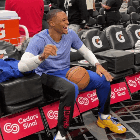Video gif. NBA player Russell Westbrook sits on the sidelines with a basketball in between his knees doing a fun shoulder dance and smile-laughing from ear to ear. 
