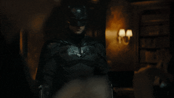 Movie gif. Robert Pattinson as Batman in "The Batman" slowly walks through a dark room and stares at us with a serious expression.