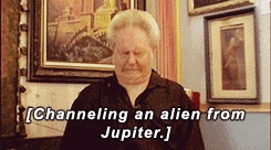 Man with brushed-up blond hair like a troll doll squeezes his eyes shut and frowns in intense concentration, slowly bobbing his head forward and backward and making an epic triple chin. Caption text attempting to explain the moment reads, “Channeling an alien from Jupiter.” Video cuts to Louis Theroux staring and smiling awkwardly at the man, maybe trying to look encouraging. 