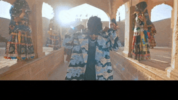 Music video gif. Badshah, wearing thick sunglasses, lifts the hood of his coat over his head in slow motion. Video then cuts to him standing with a woman singing while Badshah points at us. Text reads, “ITS YOUR BOY BADSHAH.”
