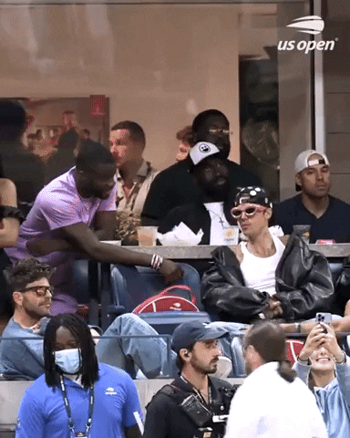 Video gif. Justin Bieber seated in the stands at the US Open wearing a leather jacket, backwards hat, and over-the-top pink oval-framed sunglasses. He high fives a man in the row behind him in a cool, casual way.