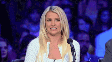 Reality TV gif. Britney Spears as a judge at a microphone. She winces and looks away uncomfortably.