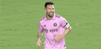 Sports gif. Soccer player Leo Messi wearing a pink Adidas jersey smiles with an open mouth, holding out his hands and running across the field toward someone.