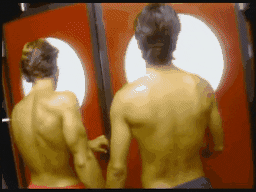 Video gif. Two men in speedos hold hands as they walk into a black tiled room together. 
