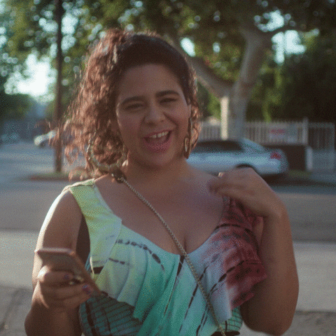TV gif. Jessica Marie Garcia as Jasmine in On My Block struts down the street while holding her phone. She scrunches her nose as she gives us an open mouth grin. She's filled with attitude and is radiating confidence as she walks.
