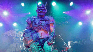 Video gif. Masked member of the band Gwar smiles and waves at us enthusiastically on stage.