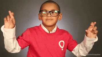 Video gif. A little boy with big glasses on his face holds his hands up and crosses his fingers. He smiles and hands his crossed fingers around. 