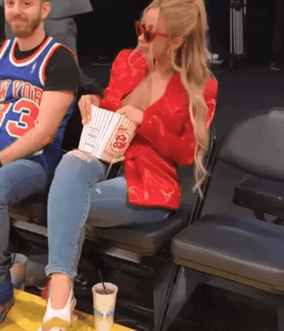 Celebrity gif. Cardi B is sitting courtside at a basketball game and is happily dancing in her seat and eating popcorn, unfazed by what's going on in front of her.