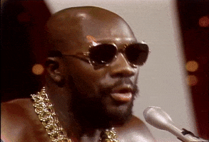 Video gif. We see Isaac Hayes close up as he sings into a microphone. We pull back to see him playing the piano, wearing an enormous gold chain and no shirt as lights twinkle behind him. 