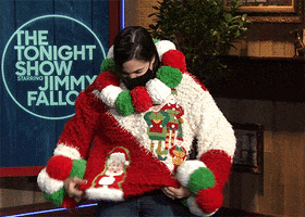 TV gif. A masked fan on The Tonight Show shows off her ugly Christmas sweater, twirling so we can see the giant red, green and white puffballs on the neck and wrists, as well as Santa and his elves on the front and a Christmas tree on the back. She finishes with a curtsy.