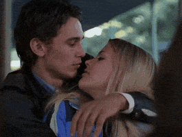 TV gif. James Franco as Daniel Desario on Freaks and Geeks hugs Busy Phillips as Kim Kelly from behind and bites her nose playfully. She pulls back from him, wincing in slight pain.