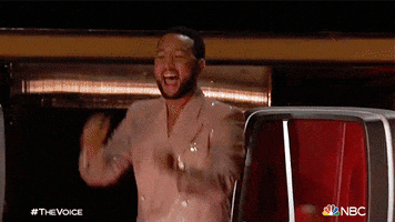 Reality TV gif. In a clip from The Voice, John Legend wears a shiny beige jacket while clapping excitedly and cheering.