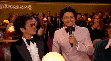 TV gif. Trevor Noah and Bruno Mars at the Grammy Awards share an awkward interaction. Trevor in a pink suit holds a microphone and grins directly at us while Bruno in a slick tuxedo and aviators stares at Noah stone-faced. Audience members around them applaud.