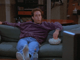 Seinfeld gif. Jerry reclining on the couch with his feet up, eating popcorn and watching TV, making a victory fist in the air