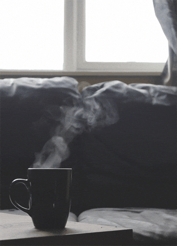 Video gif. Cozy living room scene with a coffee mug on a table, steaming, in front of a couch below a window.