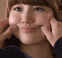 Celebrity gif. Someone squeezes the cheeks of a blinking K-pop star Bae Suzy, forcing her to smile.