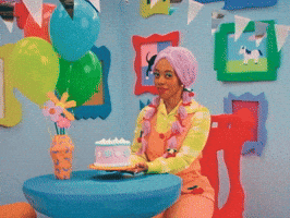 Video gif. Woman wearing a pink wig with pigtails and coral overalls sits on a chair holding a pink birthday cake in a colorful room decorated cartoon-style with bright decor including picture frames, a vase, and balloons. She smiles and says, "Happy birthday gorgeous."