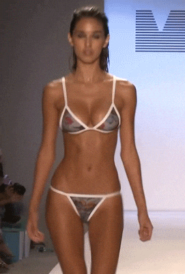Celebrity gif. Ashley Sky walks the runway modeling a bikini, boobs jiggling. 