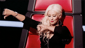 Reality TV gif. Christina Aguilera as a judge on The Voice sits in a red high backed chair. She holds both of her arms out and gives two thumbs down as she mouths the text below, "Booo!"