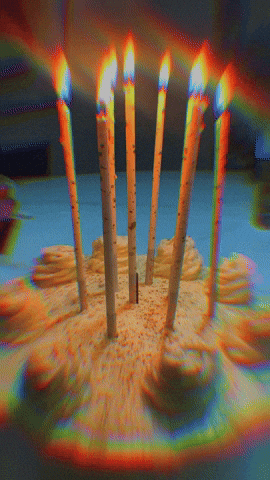 Video gif. Closeup of a birthday cake with tall candles, casting trippy beams of rainbow light across the frame. 