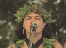 Video gif. Haunani-Kay Trask, dressed in traditional hula regalia, speaks into a microphone "Kūʻē! Kūʻē!"