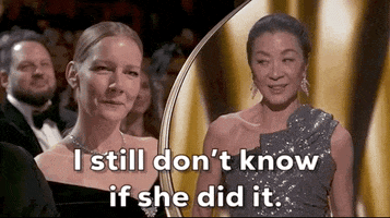 Oscars 2024 GIF. Split screen of Michelle Yeoh shaking her head in disbelief and glancing proudly at Sandra Hüller while saying, "I still don't know if she did it." Hüller grins and tilts her head to the side. 