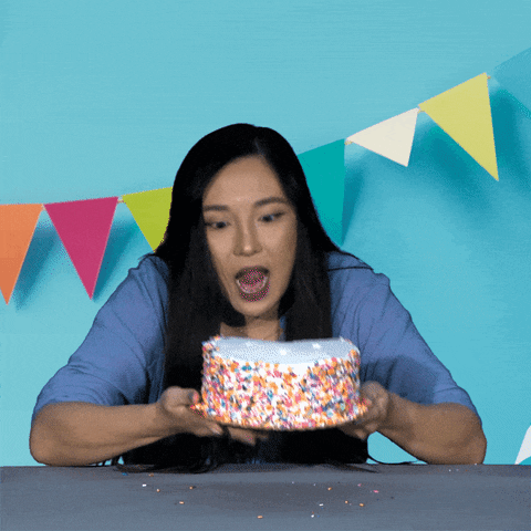Video gif. Woman leans over a table and takes a big, reckless bite into a full birthday cake, lifting her head to smack her lips, white frosting and sprinkles everywhere. 