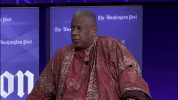 Video gif. André Leon Talley, a journalist, sits at a panel during for the Washington Post and he looks away while pursuing his lips and heavily shrugging his shoulder.