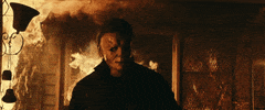 Movie gif. James Jude Courtney as Michael Myers in "Halloween Kills," walks toward us, away from a fire that rages out of a doorway behind him.
