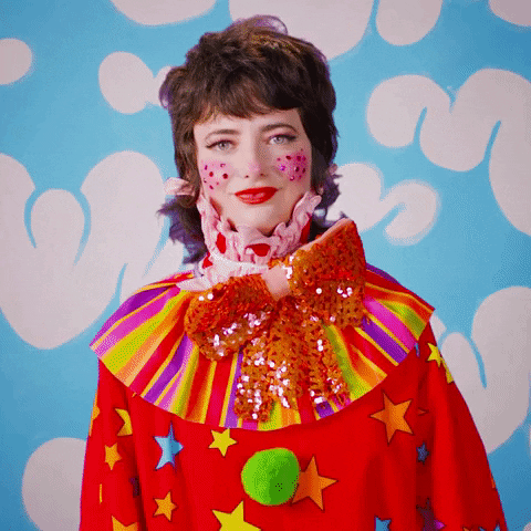 Celebrity gif. Sarah Squirm is dressed in a clown suit and she has bedazzled cheeks. She gives us a double thumbs up and big smile.
