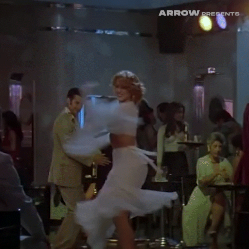 Video gif. 80s-era clip of a woman wearing a white two-piece dress with a flowing skirt and sleeves whirls and twirls around a nightclub.