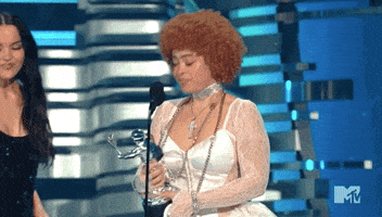 Video gif. Ice Spice at the 2023 VMAs, holding an award on stage in front of a microphone, wiping tears away and holding a hand in front of her mouth in happy disbelief. 