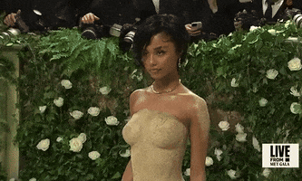 Met Gala 2024 gif. Camera zooms out from Tyla wearing a sand-colored sculpted Balmain mermaid-style dress with shimmery sand makeup on her shoulder. The dress is fitted all the way through her ankles and then pours out into a circular train behind her. She's holding an hourglass in her hand filled with sand. 
