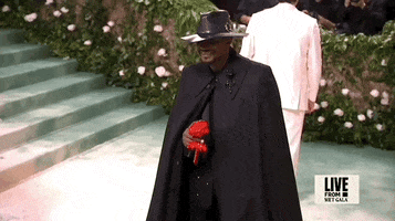 Met Gala 2024 gif. Usher, wearing an all-black custom Alexander McQueen ensemble of a cape, leather fedora, and red paper flower, lifts it to nose as if to smell it.