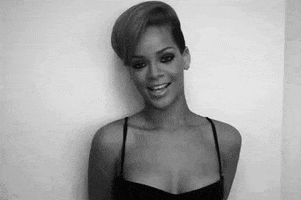 Celebrity gif. Rihanna shyly leans against a wall and smiles flirtatiously as she looks at us.