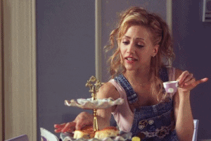 Movie gif. Brittany Murphy as Molly in Uptown Girls. She's at high tea and she holds a teacup daintily in one hand while reaching for a sandwich with the other. She takes a fat bite of the sandwich and struggles to chew.