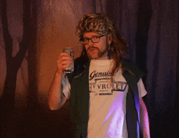 Video gif. A man wearing a mullet wig that goes past his shoulders stands in front of a creepy forest backdrop and crushes a soda can in his hand as he looks at us. 