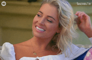 Reality TV gif. A woman from The Bachelor AU twirls her hair in a hand and tries to give a smile but it quickly turns into a thin lip of displeasure.