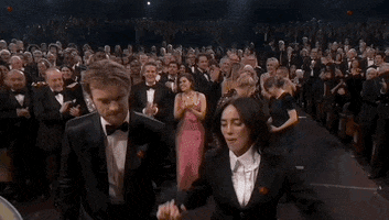 Oscars 2024 GIF. Finneas O'Connell helps Billie Eilish walk onstage while her mouth drops excitedly. She runs up to hug Ariana Grande, who presents her the award in her oversized, puffy pink dress. 