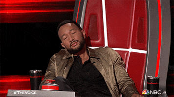 Reality TV gif. John Legend on The Voice reclines over the side of his judges chair, pretending to sleep.