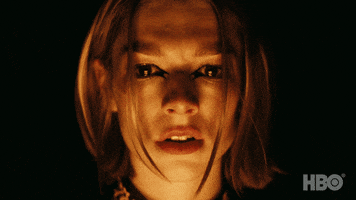 TV gif. Hunter Schafer as Jules Vaughn in Euphoria blinks as a flickering golden light gently illuminates her face and eyes in the darkness.