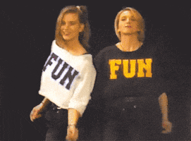 Video gif. Two women wearing shirts with the word, "FUN," on them are dancing and grooving while smiling at us.